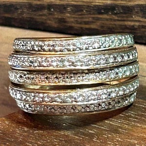 May include: A multi-band ring featuring four distinct, stacked gold-tone bands. Each band has a textured, shimmering surface with a granular, silver-toned finish that catches the light, creating a layered appearance across the entire ring structure.