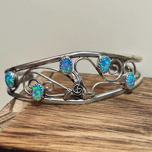 Pode incluir: Pulseira de punho prateada com seis pedras ovais de opala azul-esverdeada. A pulseira tem um design aberto com redemoinhos decorativos e um detalhe de pena. Apresentada sobre uma superfície de madeira.