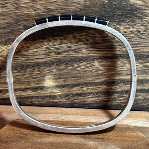 May include: A silver-toned hinged bangle bracelet with a rectangular black stone design across the top. The bracelet is displayed on a wooden surface, showcasing its polished metal and dark stone accents. The bracelet is a piece of jewelry.