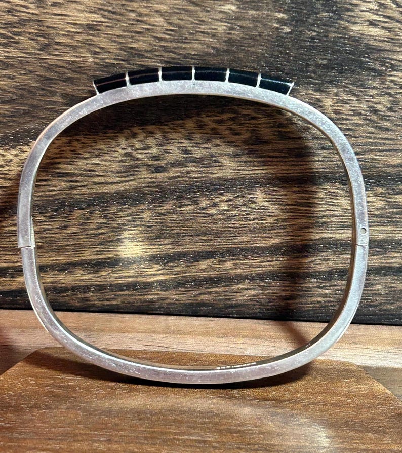 May include: A silver-toned hinged bangle bracelet with a rectangular black stone design across the top. The bracelet is displayed on a wooden surface, showcasing its polished metal and dark stone accents. The bracelet is a piece of jewelry.