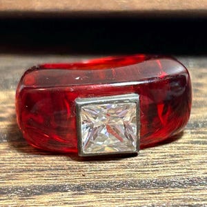Puede incluir: Un anillo de resina roja con una piedra cuadrada transparente engastada en un engaste plateado.