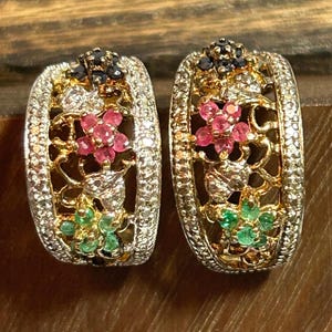 May include: A pair of gold-coloured hoop earrings with detailed floral patterns. The earrings are adorned with pink, green, and black gemstones, and a border of small, clear stones. The floral elements are set against a filigree background.
