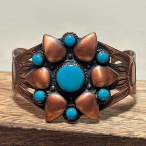 Pode incluir: Pulseira de punho de cobre com uma pedra turquesa central, rodeada por formas de pétalas de cobre e pedras turquesas menores. A pulseira tem um design aberto com detalhes gravados, em estilo sudoeste.