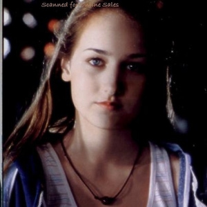 May include: A person with long brown hair and blue eyes wearing a white tank top with thin horizontal stripes in light blue and red, layered under a dark blue zip-up hoodie. A simple dark cord necklace with a single dark bead rests on the neck.