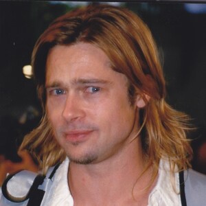 May include: A person with shoulder-length light brown hair and blue eyes wearing a white button-down shirt with a subtle geometric pattern and a silver-toned strap across the shoulder. The shirt features a collar and a partially open front neckline.