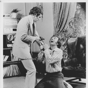 May include: A black and white still from the movie "Deadly Honeymoon" featuring two men. One man is holding a gun to the other man's throat. The man with the gun is wearing a white suit jacket and a white shirt with a dark tie. The man with the gun is standing and the other man is kneeling on the floor. The background is a living room with a couch and a record player.