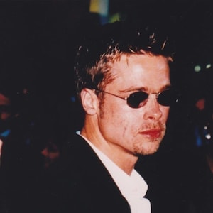 May include: A person with short, styled brown hair wearing dark, oval-shaped sunglasses with thin metal frames. The individual is dressed in a white collared shirt layered under a black jacket with a visible front zipper. The lighting is dim.