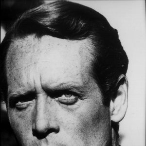 May include: Black and white portrait photograph of a man with short, dark hair. He is looking directly at the camera with a serious expression. The man has a strong jawline and a prominent nose.