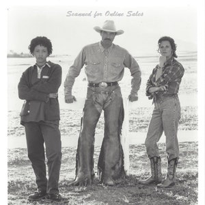 May include: A black and white promotional photograph featuring three individuals standing in a field. The central figure wears a cowboy hat, button-down shirt, and chaps. The individuals on the left and right wear casual western-style clothing.