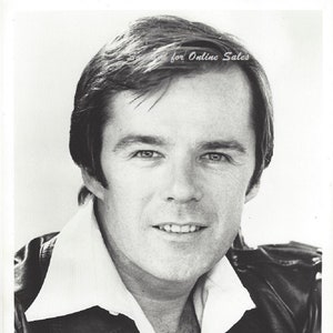May include: A black and white headshot of a person with dark hair, wearing a white collared shirt under a dark jacket. The text at the bottom reads: CEASE FIRE, shot on locations in Florida, stars Don Johnson, Lisa Blount and Robert F. Lyons.