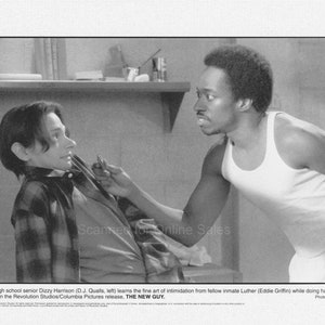 May include: A black and white photo of two men in a prison setting. The man on the left is wearing a plaid shirt and the man on the right is wearing a white tank top. The man on the right is holding a small object in his hand and is looking at the man on the left.