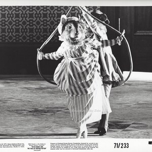 May include: A black and white photo of a person in a striped dress and a rabbit costume holding a hoop. The person is performing on a stage with a patterned floor. The photo is from the movie "Peter Rabbit and the Tales of Beatrix Potter".