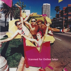 May include: A group of four people emerging from a large, torn-open magenta and yellow cardboard box on a city street. The individuals are wearing swimwear, sunglasses, and accessories, with one person wearing a pink bikini top and red sneakers.
