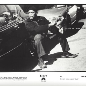 May include: A black and white photo of a man wearing a black hat and jacket, sitting next to a car. He is holding a gun in his right hand. The text "SHAFT" is printed at the bottom of the photo.