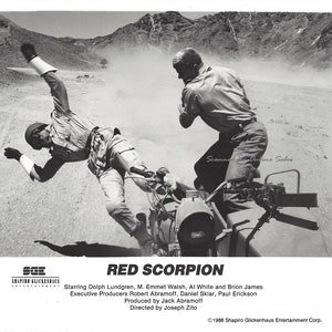 May include: A black and white photo of a scene from the action film "Red Scorpion".  Two men are in the desert, one is on a motorcycle and the other is standing behind him.  The motorcycle is flipped over and the man on the motorcycle is lying on the ground.