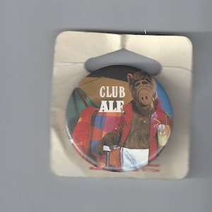 May include: A round pinback button featuring a brown, furry creature wearing a red jacket and white shorts, holding a drink. The text CLUB ALF is printed in white above the character. The button is attached to a white cardboard backing card.