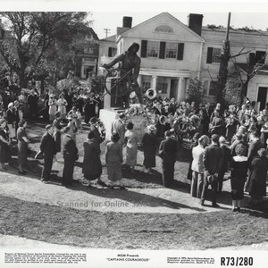 May include: A black and white photograph showing a crowd of people gathered around a statue of a person holding a ship's wheel. The statue stands on a stone base in front of a large house. The text "Scanned for Online Sales" is visible.