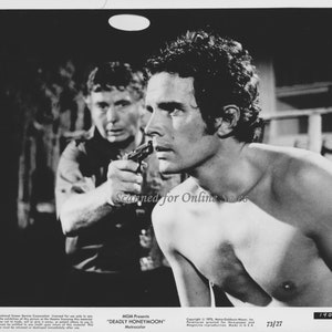 May include: A black and white photo of a man being held at gunpoint by another man. The man being held at gunpoint is shirtless and has his back to the camera. The man holding the gun is wearing a light-colored shirt. The text "MGM Presents "DEADLY HONEYMOON" Metrocolor" is visible at the bottom of the photo.