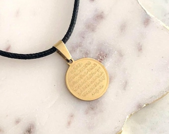 Può includere: Una collana con pendente color oro con cordino nero. Il pendente rotondo presenta testo inciso. La collana è esposta su una superficie di marmo bianco e beige. Il pendente ha un diametro di circa 2,5 cm.