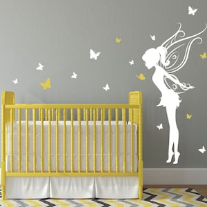 以下が含まれることがあります： 灰色の壁に、羽根と蝶のついた妖精の白いシルエット。手前に、白いシーツを敷いた黄色のベビーベッドがある。