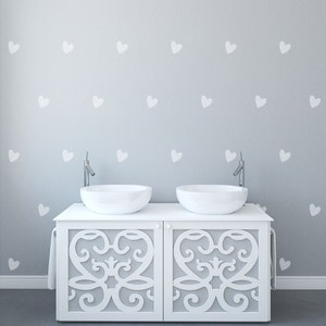 May include: A white vanity with two round sinks and chrome faucets. The vanity has decorative cutouts on the doors and a towel bar. The gray wall is decorated with white heart-shaped decals. The floor is gray.