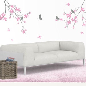 May include: A modern living room scene featuring a white sofa, a pink shag rug, and a gray wicker basket. The wall is decorated with gray branches, pink flowers, and gray birds. A white floor lamp and a white vase with branches complete the decor.