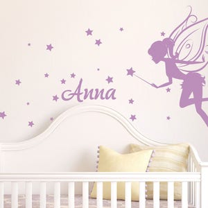 May include: A purple fairy silhouette with wings and a wand, flying above a white cot with a purple name decal that says "Anna" and a scattering of purple stars.