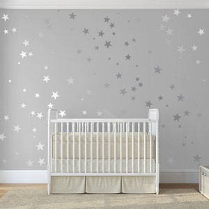 May include: A white cot with a white mattress and a white sheet in a nursery with a grey wall covered in white star decals.
