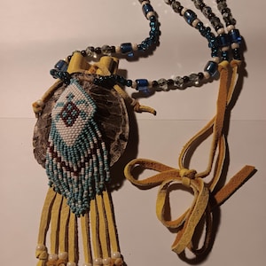 May include: A necklace featuring a circular brown textured shell pendant adorned with a diamond-shaped beaded pattern in white, light blue, and dark red. The pendant has yellow leather fringe with small beads and a strap of blue and clear beads.
