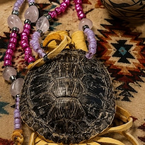 May include: A turtle shell amulet featuring a dark, textured carapace attached to light tan leather fringe. The piece is adorned with a beaded necklace strand composed of translucent light purple beads, polished pink quartz stones, and metallic silver spacers.