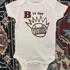 May include: White baby onesie with brown text that reads "B is for" and a cartoon illustration of a brown monster with a crown of spikes.