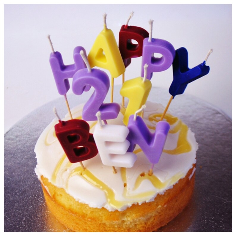 K&ouml;nnte beinhalten: Ein kleiner runder Kuchen mit wei&szlig;em Zuckerguss und gelbem Guss. Bunte Kerzen schreiben "HAPPY 27 BEN" in Lila, Gelb, Rot, Wei&szlig; und Blau. Die Kerzen sind in den Kuchen gesteckt und bilden eine festliche Geburtstagsdekoration.