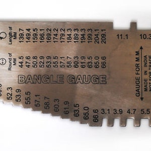 May include: A silver metal bangle gauge with measurements in millimeters and inches. The gauge is marked "BANGLE GAUGE" and "GAUGE FOR M.M." It is made in India and is not for trade.