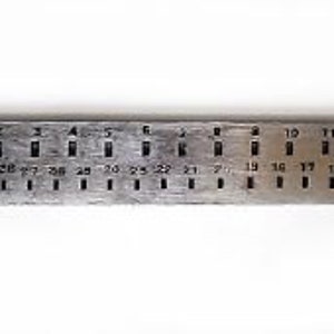 Könnte beinhalten: Ein Metalllineal mit nummerierten Löchern, beschriftet mit "STELLA" und zeigt Größen von 1 bis 15. Das Lineal hat Markierungen für verschiedene Größen in Millimetern.