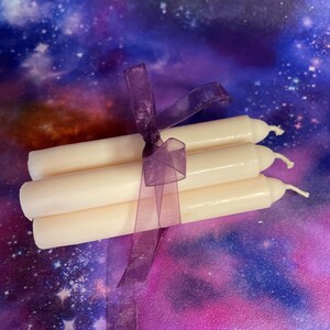 Peut inclure: Un lot de trois bougies effil&eacute;es blanc cass&eacute;, lisses, attach&eacute;es ensemble par un ruban en organza violet translucide. Chaque bougie pr&eacute;sente un sommet arrondi avec une m&egrave;che en coton visible, dispos&eacute;es horizontalement en pile.