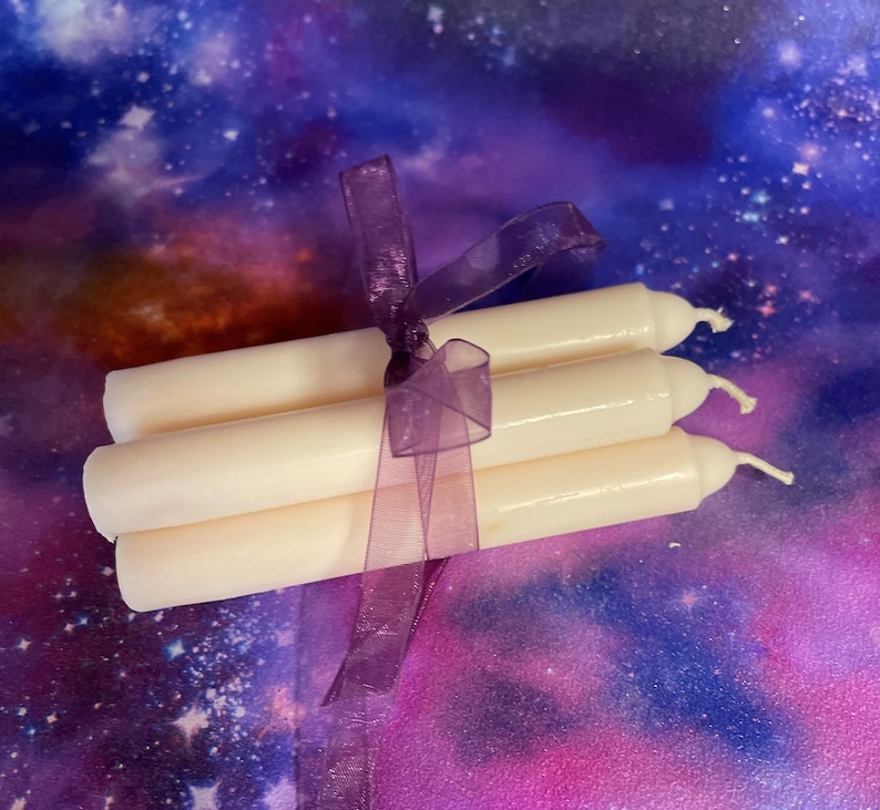 Peut inclure: Un lot de trois bougies effil&eacute;es blanc cass&eacute;, lisses, attach&eacute;es ensemble par un ruban en organza violet translucide. Chaque bougie pr&eacute;sente un sommet arrondi avec une m&egrave;che en coton visible, dispos&eacute;es horizontalement en pile.
