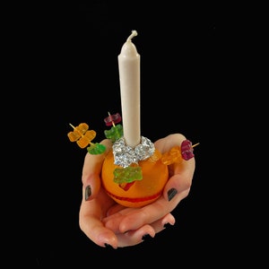 Peut inclure: Un fruit orange vif servant de base &agrave; une longue bougie blanche, fix&eacute;e avec du papier aluminium argent&eacute;. Plusieurs cure-dents en bois sont ins&eacute;r&eacute;s dans l'orange, chacun d&eacute;cor&eacute; de bonbons g&eacute;lifi&eacute;s color&eacute;s rouges, verts et jaunes.