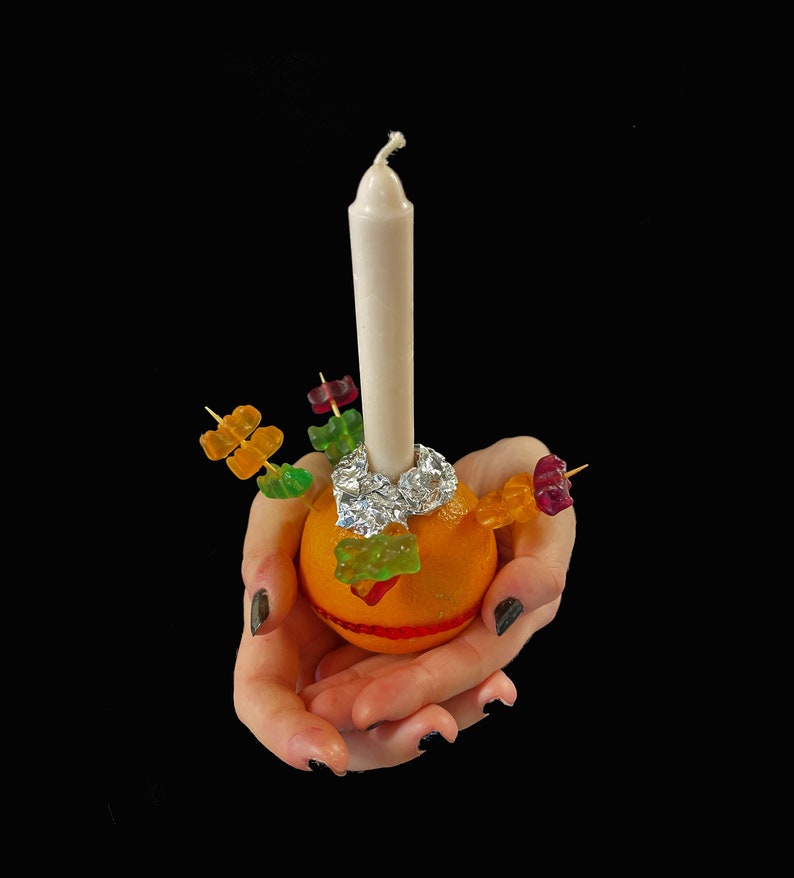 Peut inclure: Un fruit orange vif servant de base &agrave; une longue bougie blanche, fix&eacute;e avec du papier aluminium argent&eacute;. Plusieurs cure-dents en bois sont ins&eacute;r&eacute;s dans l'orange, chacun d&eacute;cor&eacute; de bonbons g&eacute;lifi&eacute;s color&eacute;s rouges, verts et jaunes.