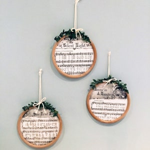 May include: Three round, wooden, framed ornaments with greenery and sheet music featuring the lyrics to the Christmas carols "Silent Night", "Away in a Manger", and "The First Noel".