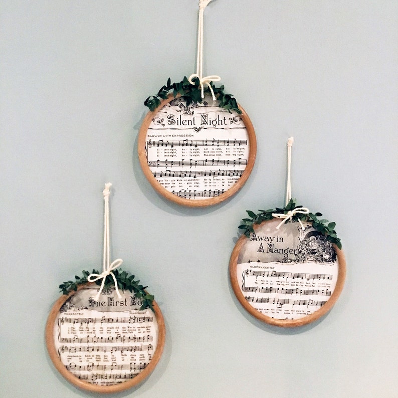 May include: Three round, wooden, framed ornaments with greenery and sheet music featuring the lyrics to the Christmas carols "Silent Night", "Away in a Manger", and "The First Noel".