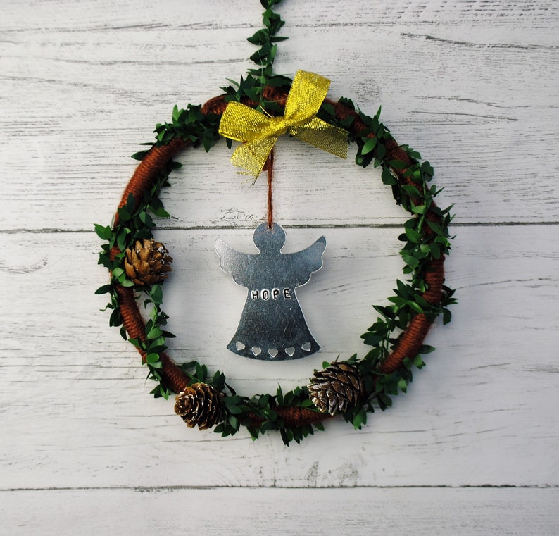 May include: A brown wreath with green leaves and a yellow bow. A silver angel ornament with the word "HOPE" hangs from the bow.