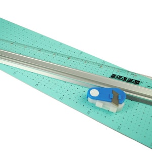 K&ouml;nnte beinhalten: Eine t&uuml;rkisfarbene Schneidematte mit einem silbernen Lineal und einem blau-wei&szlig;en Rollschneider. Das Lineal ist in Zoll unterteilt und die Schneidematte hat ein Rastermuster. Der Rollschneider hat eine wei&szlig;e Klinge und einen blauen Griff. Auf dem Lineal steht "DAFA" gedruckt.
