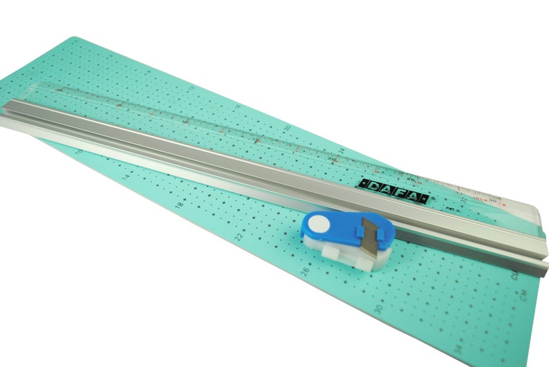 K&ouml;nnte beinhalten: Eine t&uuml;rkisfarbene Schneidematte mit einem silbernen Lineal und einem blau-wei&szlig;en Rollschneider. Das Lineal ist in Zoll unterteilt und die Schneidematte hat ein Rastermuster. Der Rollschneider hat eine wei&szlig;e Klinge und einen blauen Griff. Auf dem Lineal steht "DAFA" gedruckt.