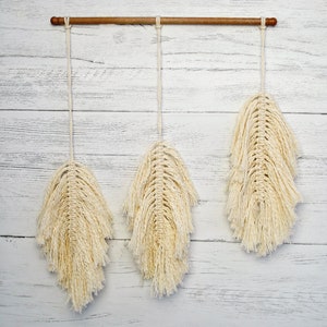 May include: Three macrame wall hangings with a wood dowel rod at the top. The hangings are made of natural cotton cord and are in the shape of feathers. The hangings are cream colored and have a fringed edge.