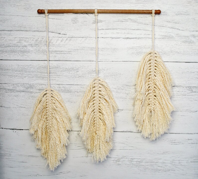 May include: Three macrame wall hangings with a wood dowel rod at the top. The hangings are made of natural cotton cord and are in the shape of feathers. The hangings are cream colored and have a fringed edge.