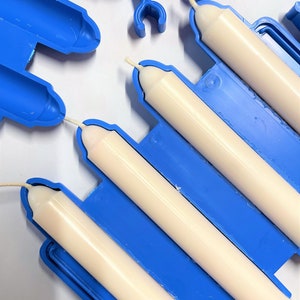 Peut inclure: Un moule &agrave; bougie en plastique bleu contenant trois bougies effil&eacute;es blanc cass&eacute; avec des m&egrave;ches visibles. Le moule est compos&eacute; de sections cylindriques reli&eacute;es avec de petits clips en plastique bleu pour fixer les deux moiti&eacute;s du moule.