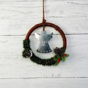 May include: A brown and silver Christmas wreath with a metal angel ornament that says "LOVE" in the center. The wreath is decorated with pine branches, pine cones, and red berries.