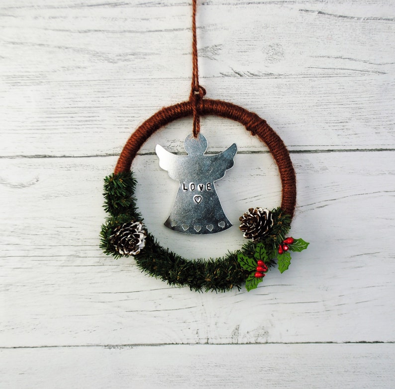 May include: A brown and silver Christmas wreath with a metal angel ornament that says "LOVE" in the center. The wreath is decorated with pine branches, pine cones, and red berries.