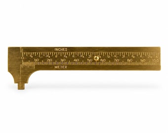 May include: A brass sliding caliper featuring dual measurement scales. The top scale is marked INCHES with increments up to 4 inches, while the bottom scale is marked METER with increments up to 100 millimeters. A small brass knob sits on the sliding arm.