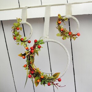 May include: Three wreaths made of woven branches with red berries and green leaves hanging from a white wall.